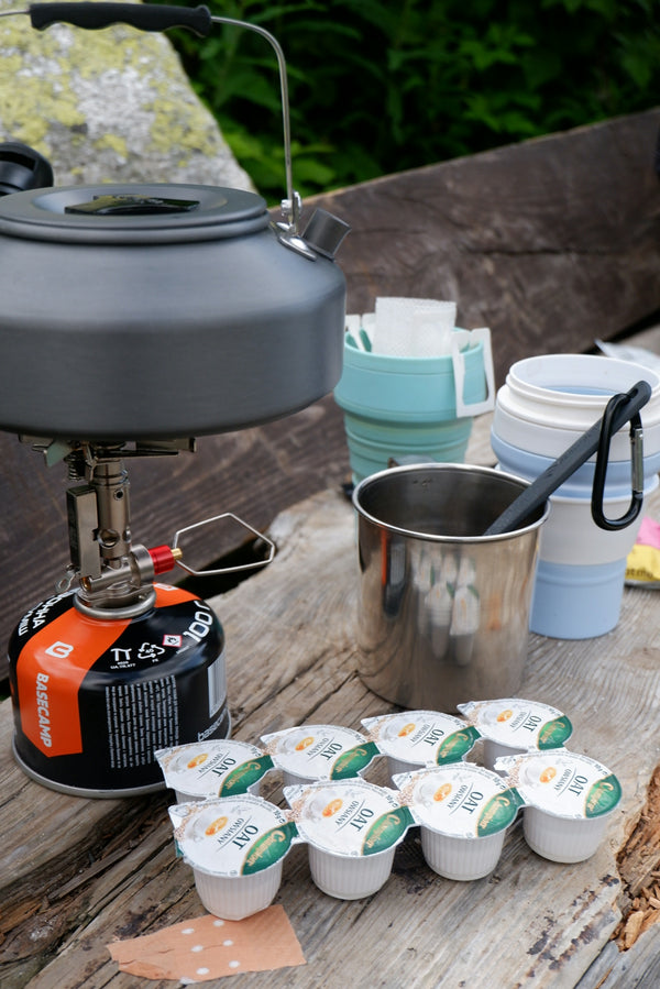 A camping stove with a kettle, metal mug, and small oat creamers on wood