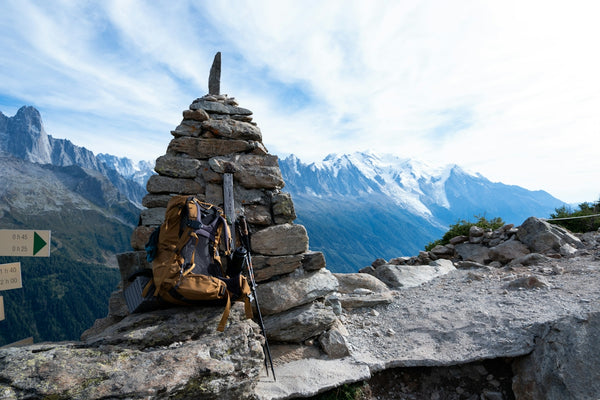 A brown hiking backpack and trekking poles leaning against a stone cairn in the mountains